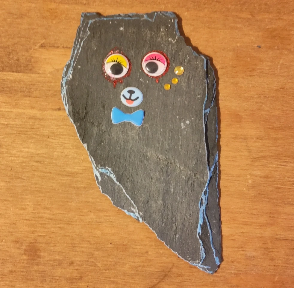 A piece of broken roof slate with a face made of googly eyes, dripping red glitter and a few other stickers stuck on it and bright metallic blue highlights around the edges.