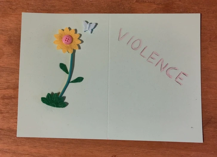 A card with a flower made of some simple craft supplies and a butterfly flying over it. Opposite the flower is a the word “violence” written in pink with black shadows.