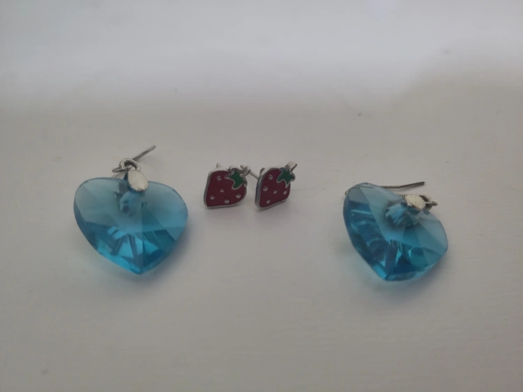 Two pairs of earrings: Studs with a strawberry design and fishhooks with blue translucent hearts.