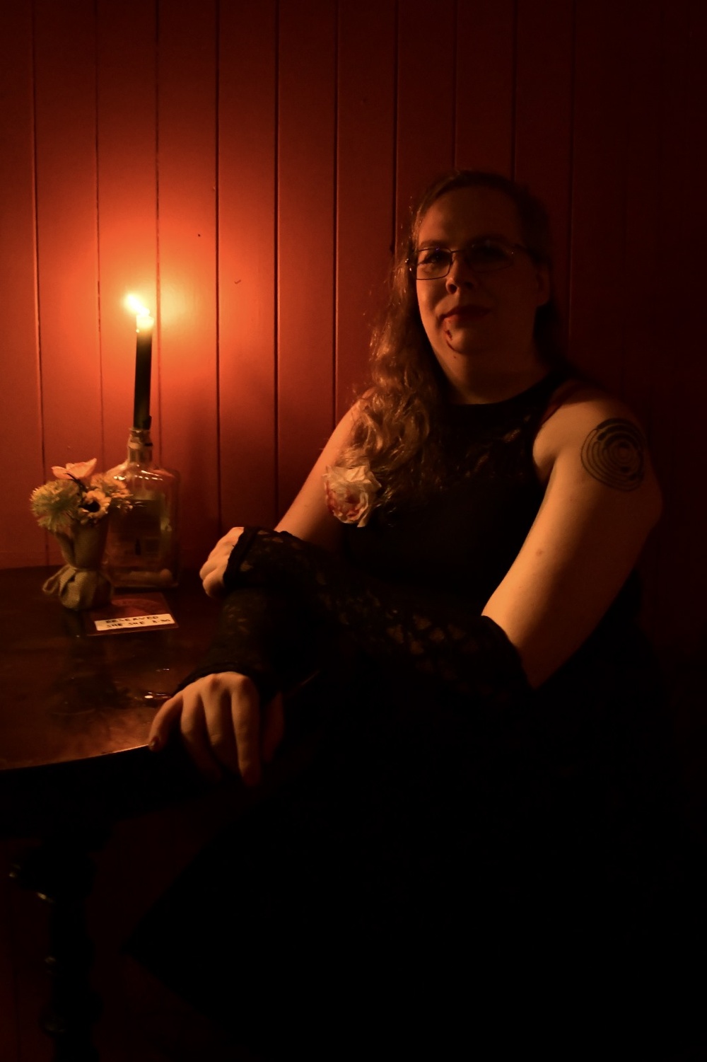 Caoimhe in a black dress and black lace gloves, sitting in deep red candlelight. There is a drop of fake blood on her chin and a red-stained white rose pinned to her dress.